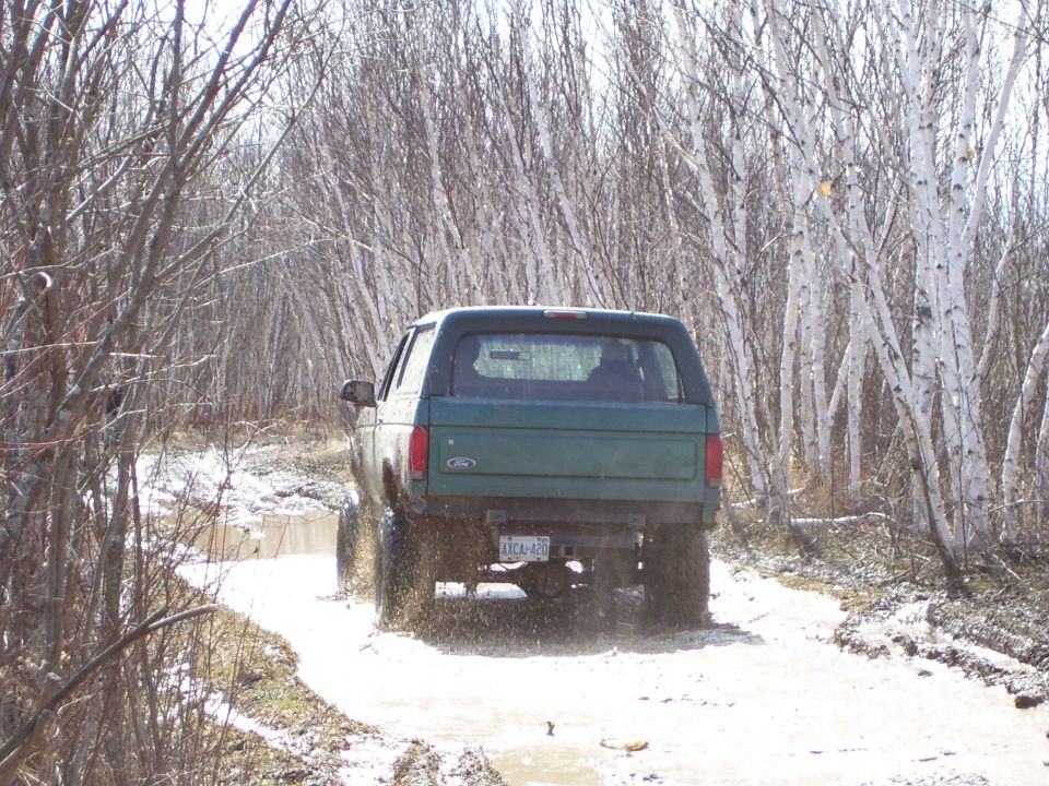 Green 1996 Ford Bronco driving through a muddy trail with trees on either side