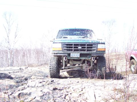 1996 Ford Bronco - A lifted green Ford Bronco is shown from the front on a rocky terrain, with another vehicle visible on the right.