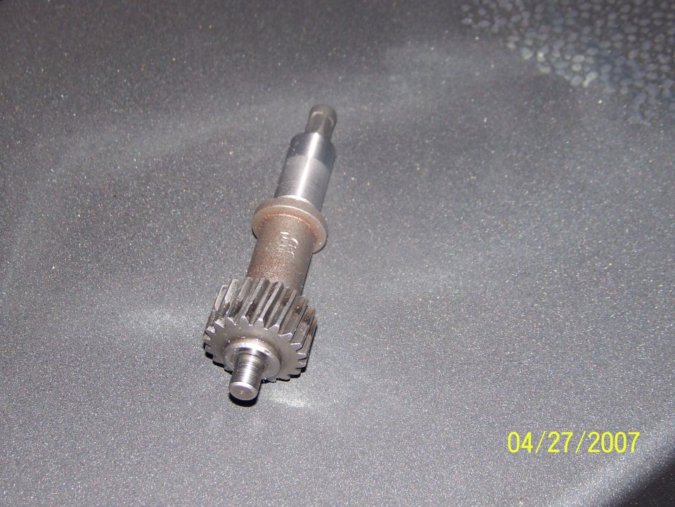 Steel shaft with small gear, a metal drivetrain part on gray surface, dated 04/27/2007
