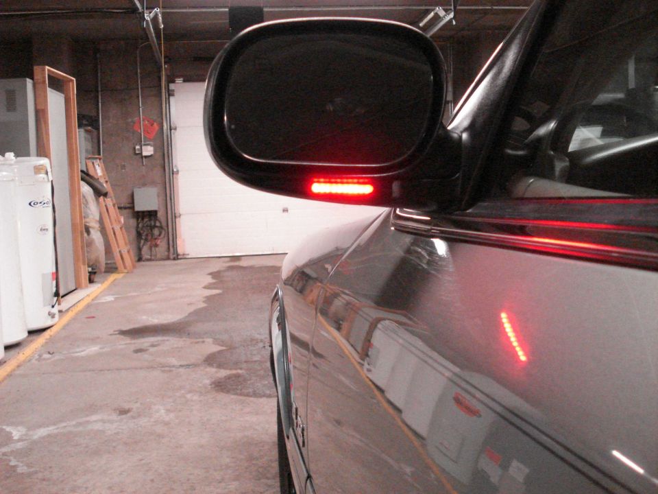 Driver-side mirror with lit red LED turn signal on gray sedan in a garage