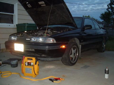 1989 Mazda MX-6 - A black 1989 Mazda MX-6 GT coupe is shown with its hood open and a jump starter connected, suggesting it needed a boost.