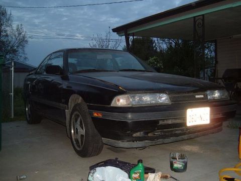 1989 Mazda MX-6 - A black 1989 Mazda MX-6 is parked in a driveway under an overhang, with a license plate visible.