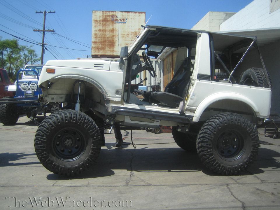 White Suzuki Samurai with large tires and lifted suspension, driver's door open, rear spare tire visible
