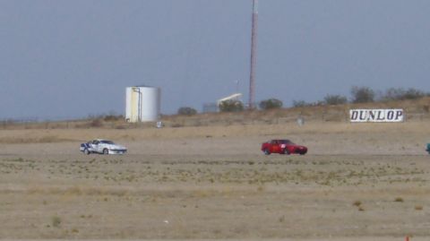 1990 Ford Probe - Two cars compete on a dusty racetrack under a hazy sky, with a water tower and advertising visible.