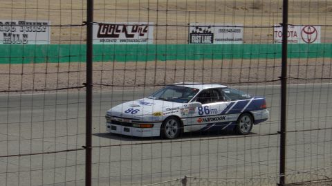 1990 Ford Probe - A white and blue race car with the number 86 is seen on a track, behind a fence.