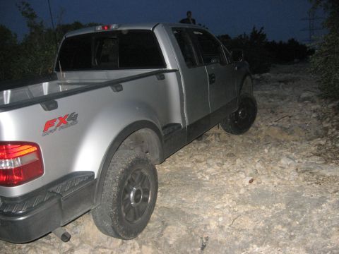 1979 Ford Bronco - A silver pickup truck is seen from the rear on a rough, rocky terrain as daylight fades.