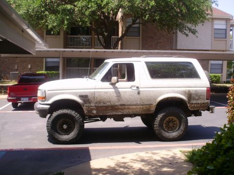 1992 Ford Bronco - A muddy white 1992 Ford Bronco is parked in a lot next to a red pickup truck.