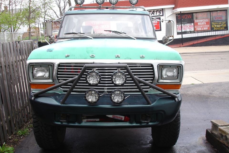 Front view of a teal Ford Bronco with black brush guard, four KC lights, and roof lights, parked by a meat market.