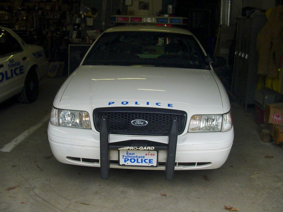 Front view of a 2002 Ford Crown Victoria police car with 'POLICE' on the hood and a light bar on top. Another police car is visible to the left