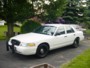 2002 Ford Crown Victoria - A white 2002 Ford Crown Victoria is parked on a driveway with trees and foliage visible.