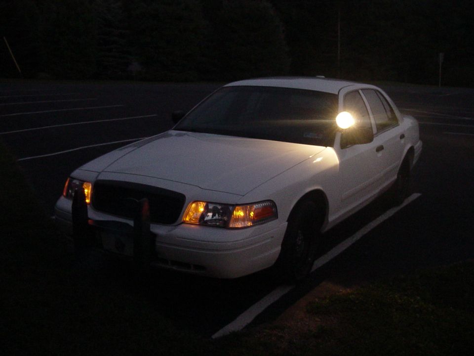 White 2002 Ford Crown Victoria with a spotlight on the driver's side mirror, parked at night