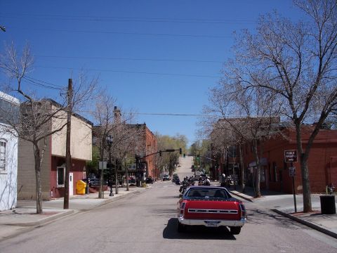 1972 Ford Ranchero - A red pickup truck leads a procession of vehicles and motorcycles down a tree-lined street on a sunny day.
