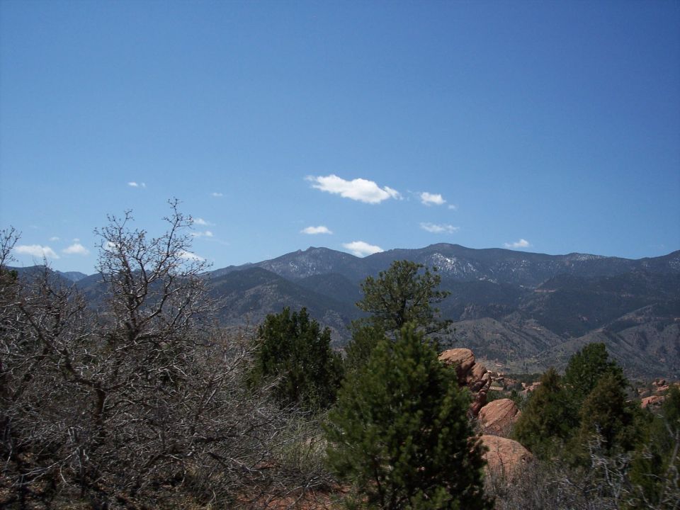 Mountain range under a blue sky with scattered clouds, foreground shows trees and red rocks
