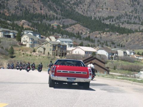 1972 Ford Ranchero - A red 1972 Ford Ranchero leads a group of motorcycles on a road with houses on a hillside.