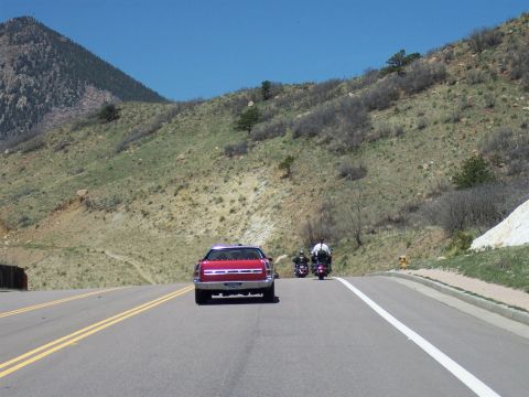 1972 Ford Ranchero - A red coupe leads two motorcycles on a highway through a mountainous landscape under a clear blue sky.
