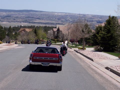 1972 Ford Ranchero - A convoy of motorcycles follows a red coupe utility vehicle through a residential neighborhood with a scenic view in the background.