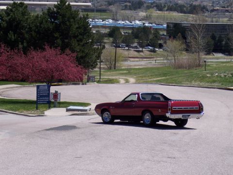 1972 Ford Ranchero - A red Ford Ranchero pickup truck is parked on a paved surface, with trees and buildings in the distance.
