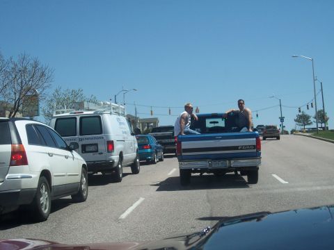 1972 Ford Ranchero - Two men ride in the bed of a blue Ford pickup truck as traffic moves along a sunny day.