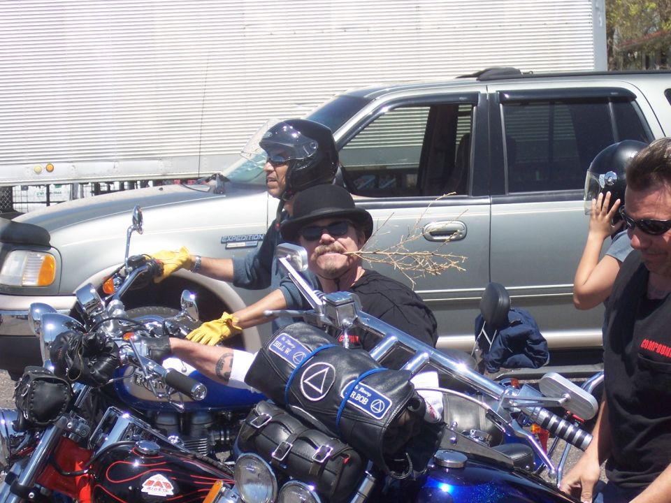 Two men on motorcycles, one wearing a hat and holding a twig in his mouth. A silver SUV is behind them
