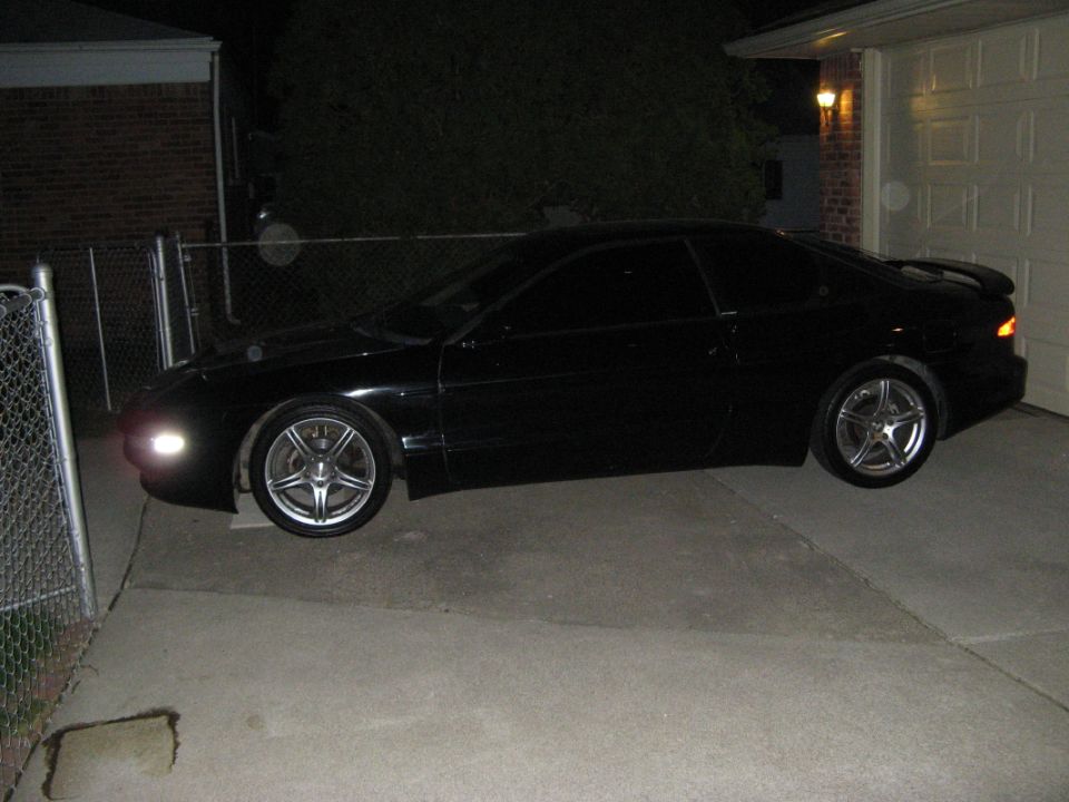 Black coupe parked in driveway at night, side view. Aftermarket wheels and spoiler visible