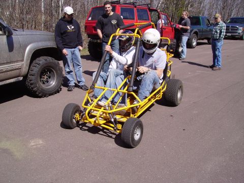 2000 Chevrolet Tahoe - A yellow go-kart with two people in helmets is parked next to a silver SUV, with other vehicles and people in the background.