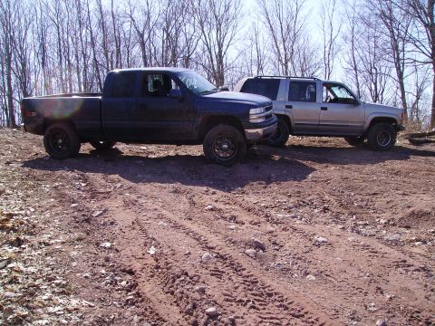 2000 Chevrolet Tahoe - Two SUVs, a Chevrolet Tahoe and a pickup truck, are parked on a dirt road with trees in the background.