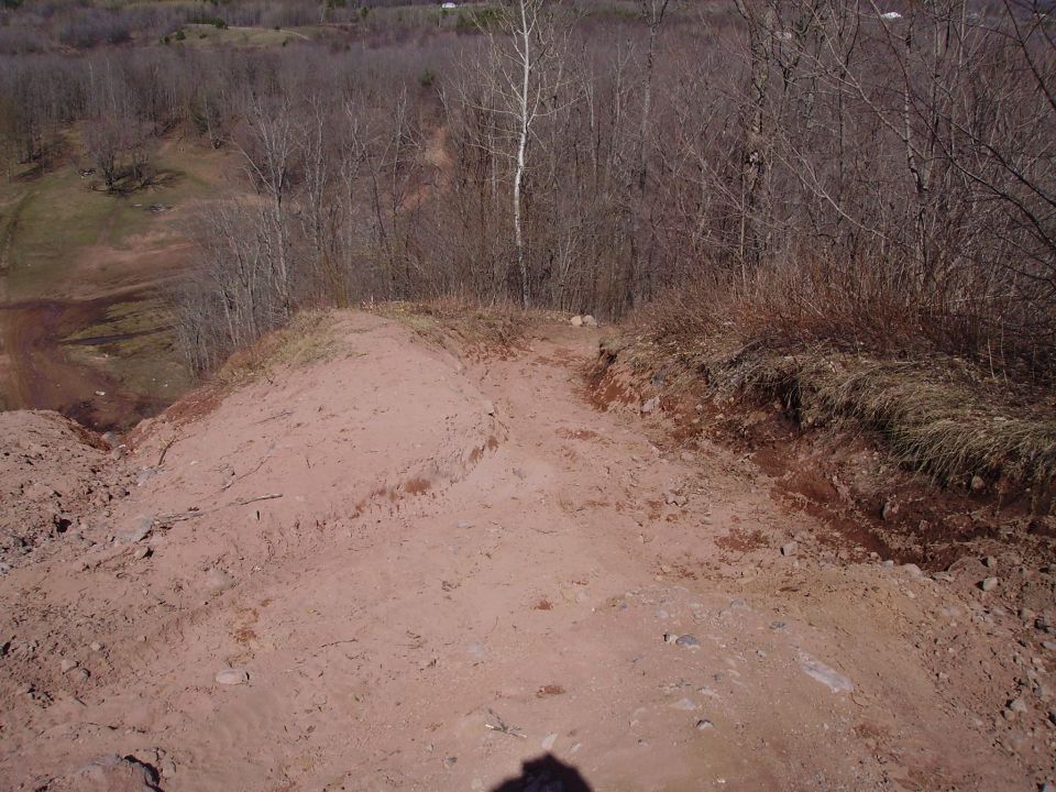 Dirt path winding down a hill with trees in the background. Supermotors logo in the corner