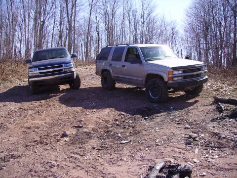 2000 Chevrolet Tahoe - Two Chevrolet SUVs are parked on a dirt path surrounded by trees. The silver SUV is in the foreground.