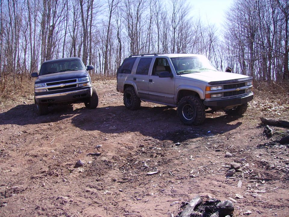 Two Chevy Tahoes parked on a dirt trail in a wooded area. The one on the right is silver, the one on the left is dark blue