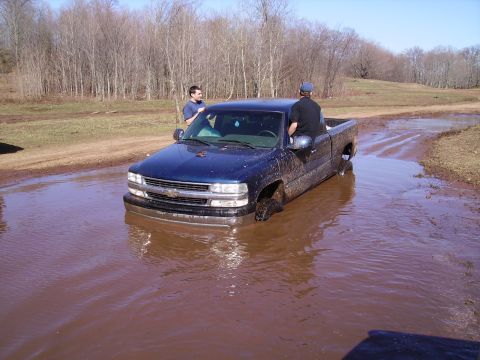 2000 Chevrolet Tahoe - A blue pickup truck is stuck in a muddy puddle with two people nearby. The grille suggests a 1999 model year.