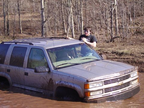 2000 Chevrolet Tahoe - A person stands by a muddy Chevrolet Suburban that is partially submerged in water, surrounded by trees.