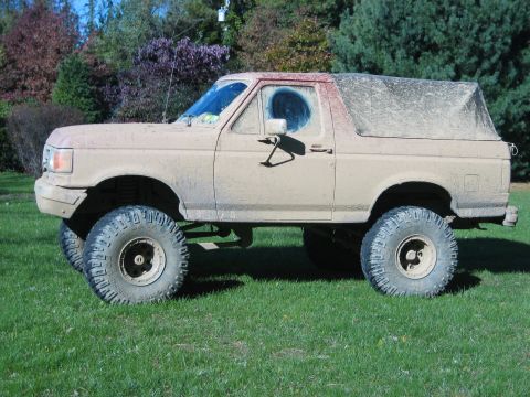 1990 Ford Bronco - Driver-side profile of a lifted Ford Bronco parked on a lawn and coated head to toe in dried tan mud, with only a wiped patch on the door g…