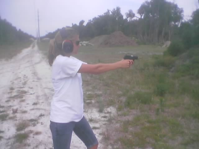Woman in white t-shirt and jeans, wearing ear protection, aims a handgun outdoors