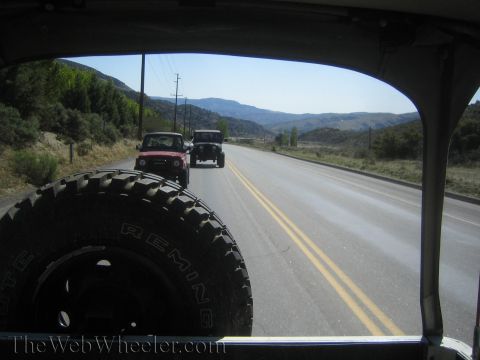 1987 Suzuki Samurai - Two off-road vehicles travel down a two-lane highway surrounded by hills and trees under a clear blue sky.