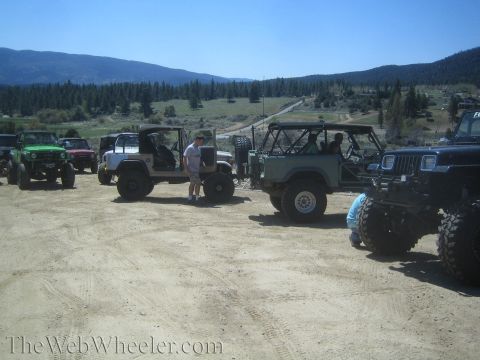 1987 Suzuki Samurai - A group of off-road vehicles are parked in a dusty lot, with a scenic mountain range and trees in the distance.