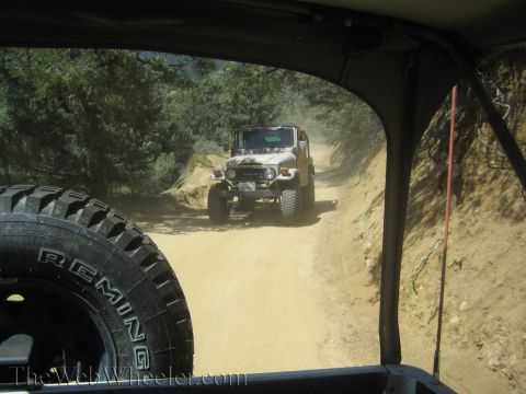 1987 Suzuki Samurai - Two off-road vehicles travel on a dusty dirt road through a wooded area.