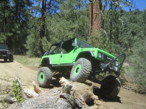 1987 Suzuki Samurai - A modified green off-road vehicle navigates a rocky trail in a wooded area, with another vehicle visible behind it.