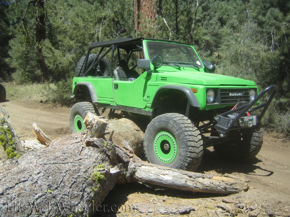 Green 1987 Suzuki Samurai with off-road tires and winch, navigating a fallen log on a dirt trail