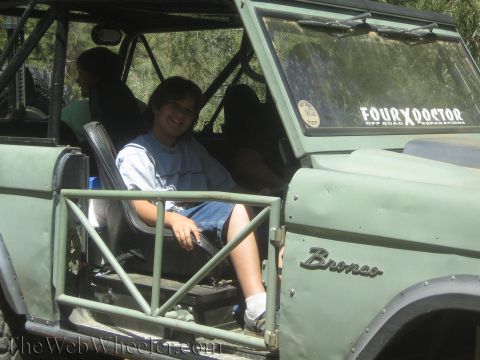 1987 Suzuki Samurai - A young person sits in the driver's seat of a green Ford Bronco, which has a roll cage and off-road preparations sticker on the windshield.