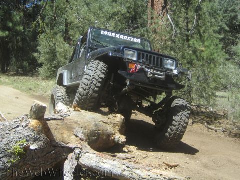 1987 Suzuki Samurai - A black Jeep Wrangler navigates a challenging off-road course, climbing over a fallen log on a dirt trail.