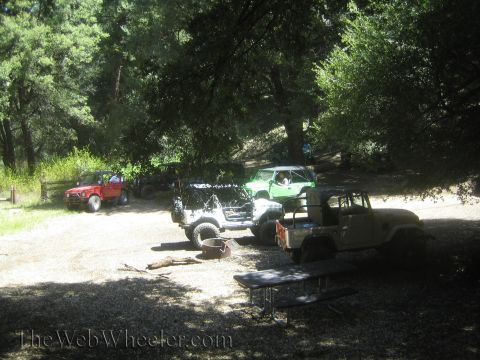 1987 Suzuki Samurai - Multiple off-road vehicles are parked in a sun-dappled wooded area.