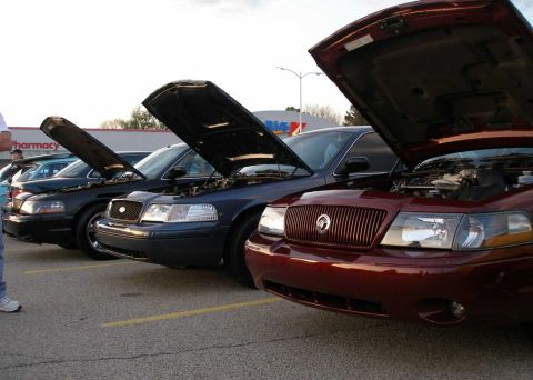 1998 Ford Crown Victoria - A line of cars, including a maroon Mercury Grand Marquis with its hood open, parked in a lot.