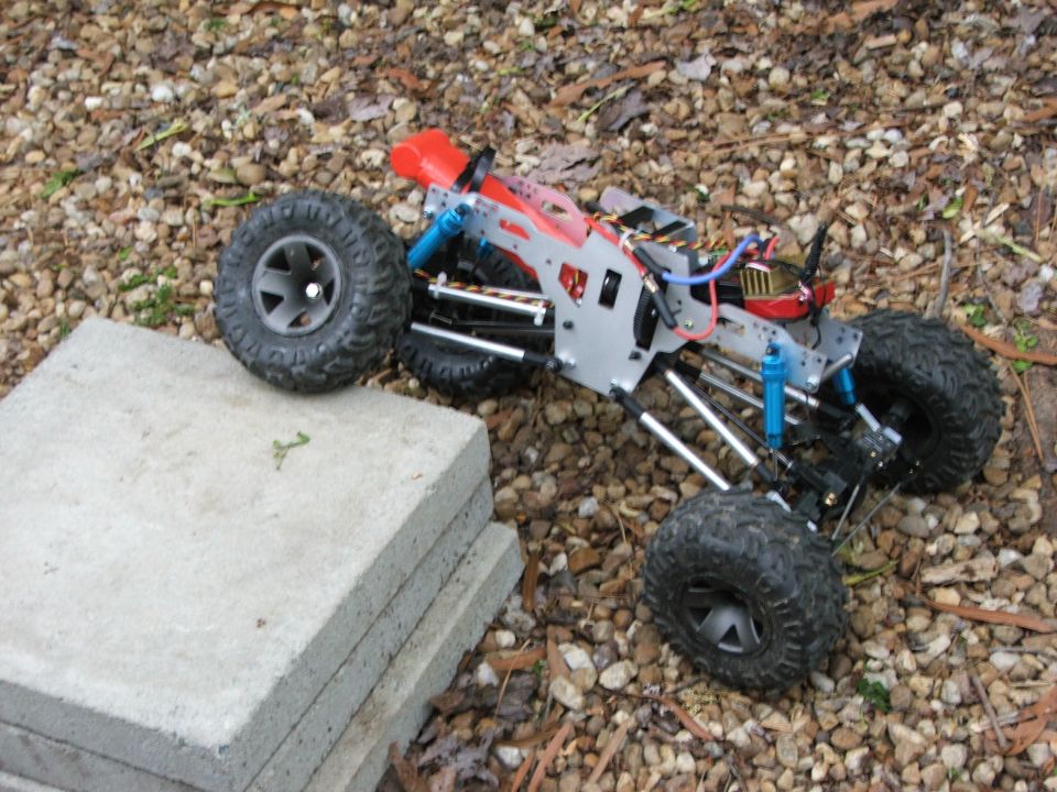 RC rock crawler chassis with large tires, blue shocks, and red bodywork, resting on gravel and concrete slabs