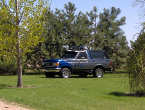 1987 Ford Bronco - A two-tone Ford Bronco sits on a grassy lawn, surrounded by trees under a clear sky.