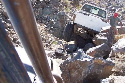1990 Ford Bronco - A white pickup truck navigates a challenging rocky trail, with another vehicle's roll cage visible in the foreground.