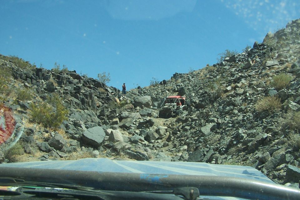 Two off-road vehicles on a steep, rocky trail under a clear blue sky. One vehicle is red, the other is partially visible
