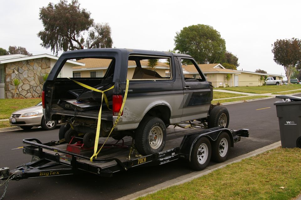 Ford Bronco on a Big Tex trailer, strapped down. The rear section is open, showing some interior components. A silver sedan is parked on the street