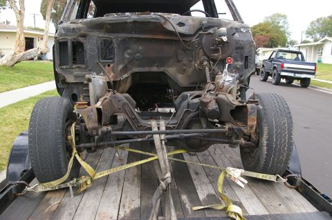 1986 Ford Bronco - The front end of a vehicle, stripped down and on a trailer, with a blue pickup truck visible in the distance.