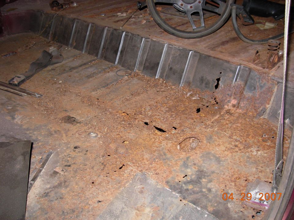 Interior of a rusty vehicle with holes in the floor and a steering wheel visible