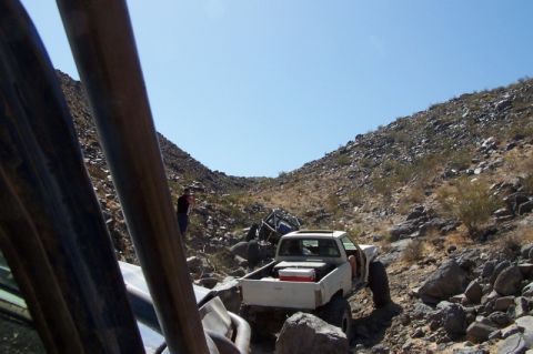 1990 Ford Bronco - Two off-road vehicles traverse a , rocky terrain under a clear blue sky.